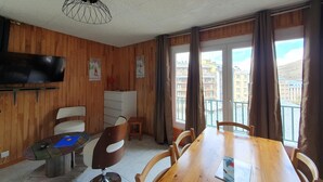 Ein Zimmer mit holzgetäfelten Wänden, einem Holzesstisch mit Stühlen, einem kleinen weißen Schrank, einem an der Wand montierten Fernseher und einem Fenster mit Vorhängen.