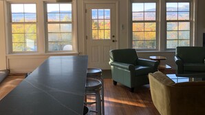 A spacious living area with a long table, multiple chairs, and large windows offering a view of autumn foliage. - Mid Century Modern Panoramic Mountain View House (New Paltz)