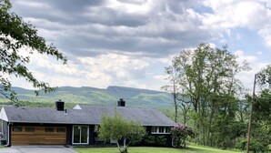 A modern house with a garage, a front yard, and a view of green hills in the background. - Mid Century Modern Panoramic Mountain View House (New Paltz)
