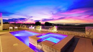 Una piscina con borde de piedra, un muro de piedra y vistas de la ciudad al anochecer.