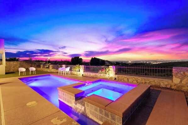 Une piscine bordée de pierres, un mur en pierre et une vue sur la ville au crépuscule.