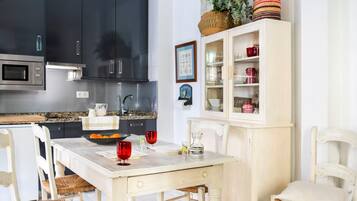 Een moderne keuken met donkere kasten, een witte eettafel, rieten stoelen, en een kast met glazen deuren waar keukengerei in staat.