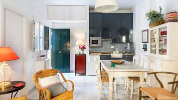 Een moderne keuken met donkere kastjes, een witte eettafel, rieten stoelen en een heldere hanglamp.