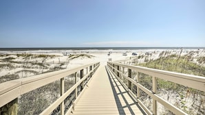 En træbro der fører til en strand med klitter og en klar himmel.