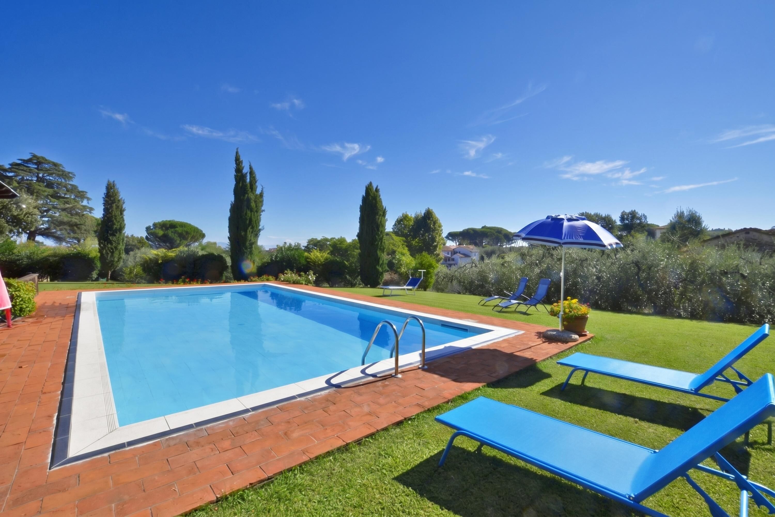 Ein rechteckiger Pool mit Backsteinrand, umgeben von einer Rasenfläche und blauen Liegestühlen.