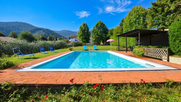 A rectangular swimming pool with a clear blue water surface, surrounded by a brick deck. There are blue lounge chairs and a covered seating area with a red umbrella. - Casa Andreini, Capannori, Italy (Capannori)
