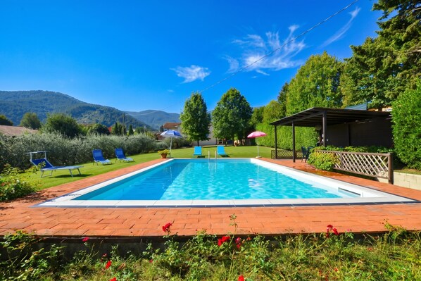 A rectangular swimming pool with a clear blue water surface, surrounded by a brick deck. There are blue lounge chairs and a covered seating area with a red umbrella. - Casa Andreini, Capannori, Italy (Capannori)