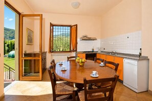 A kitchen with a dining table, chairs, a window with curtains, a view of greenery, and a white dishwasher. - Casa Andreini, Capannori, Italy (Capannori)