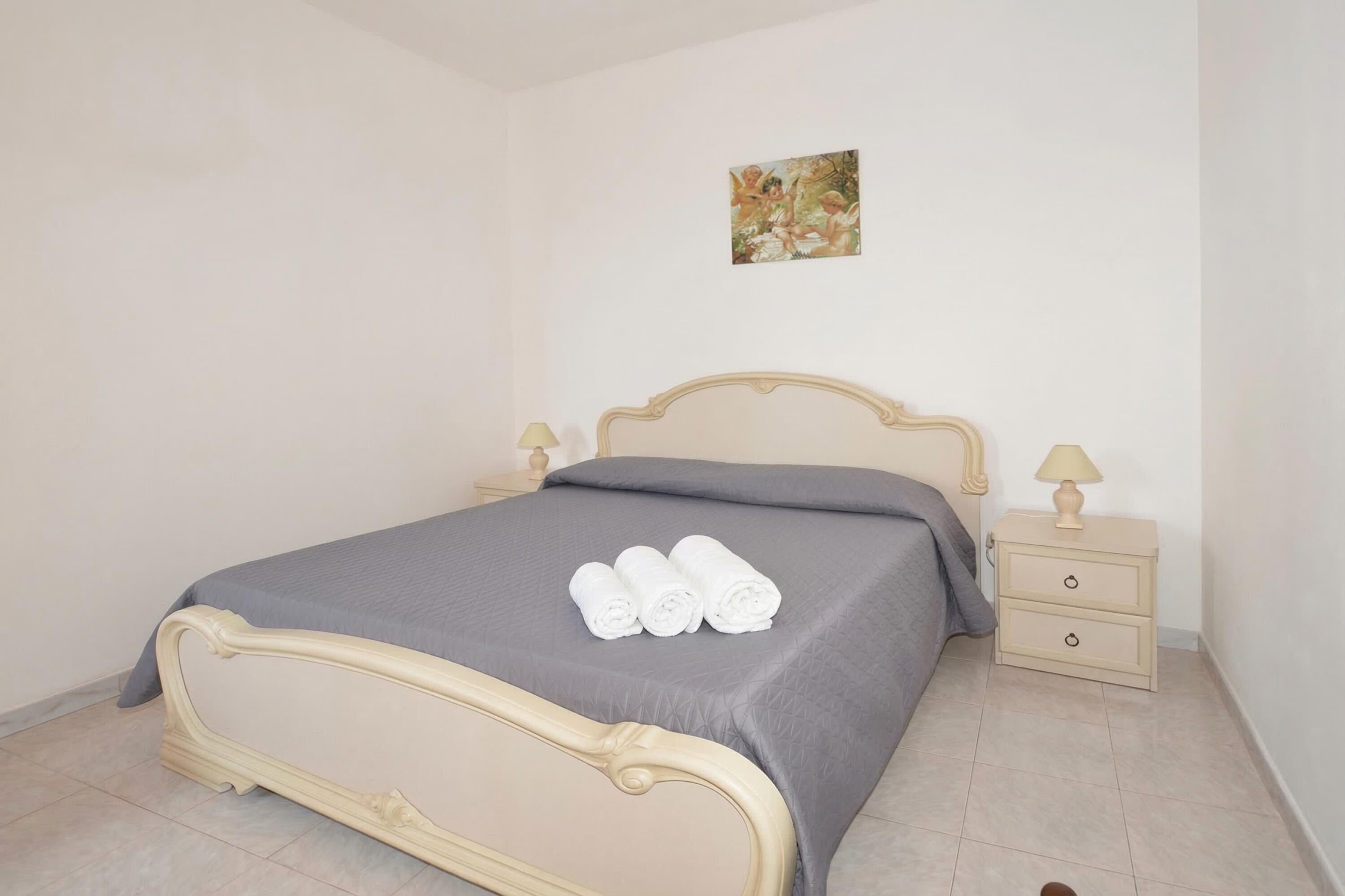 A neatly made bed with a grey bedspread and three rolled towels, a bedside table with a lamp, and a small painting on the wall.
