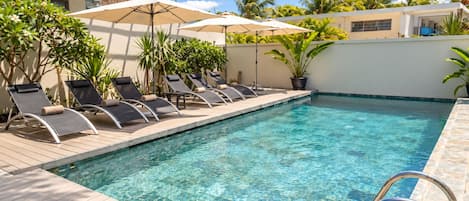 A clear swimming pool with lounge chairs and umbrellas, surrounded by greenery and a modern building.