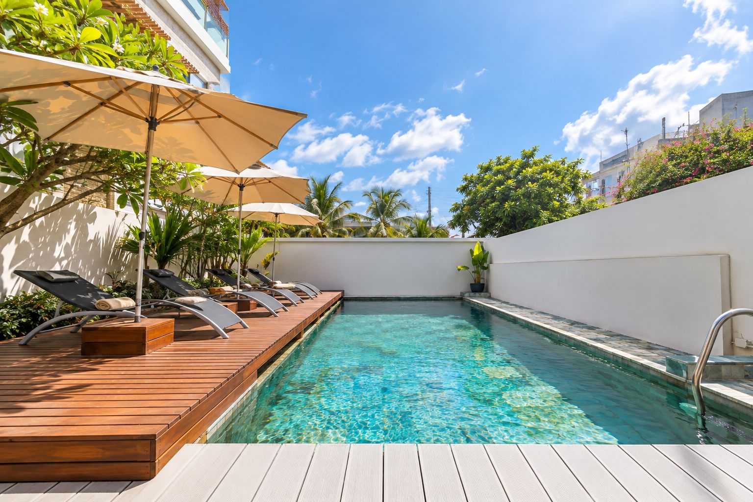A swimming pool with wooden deck and lounge chairs, surrounded by greenery and a white wall.
