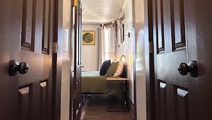 Un pasillo estrecho con puertas de madera a ambos lados, que conduce a un dormitorio con cama, almohadas y una planta colgante.