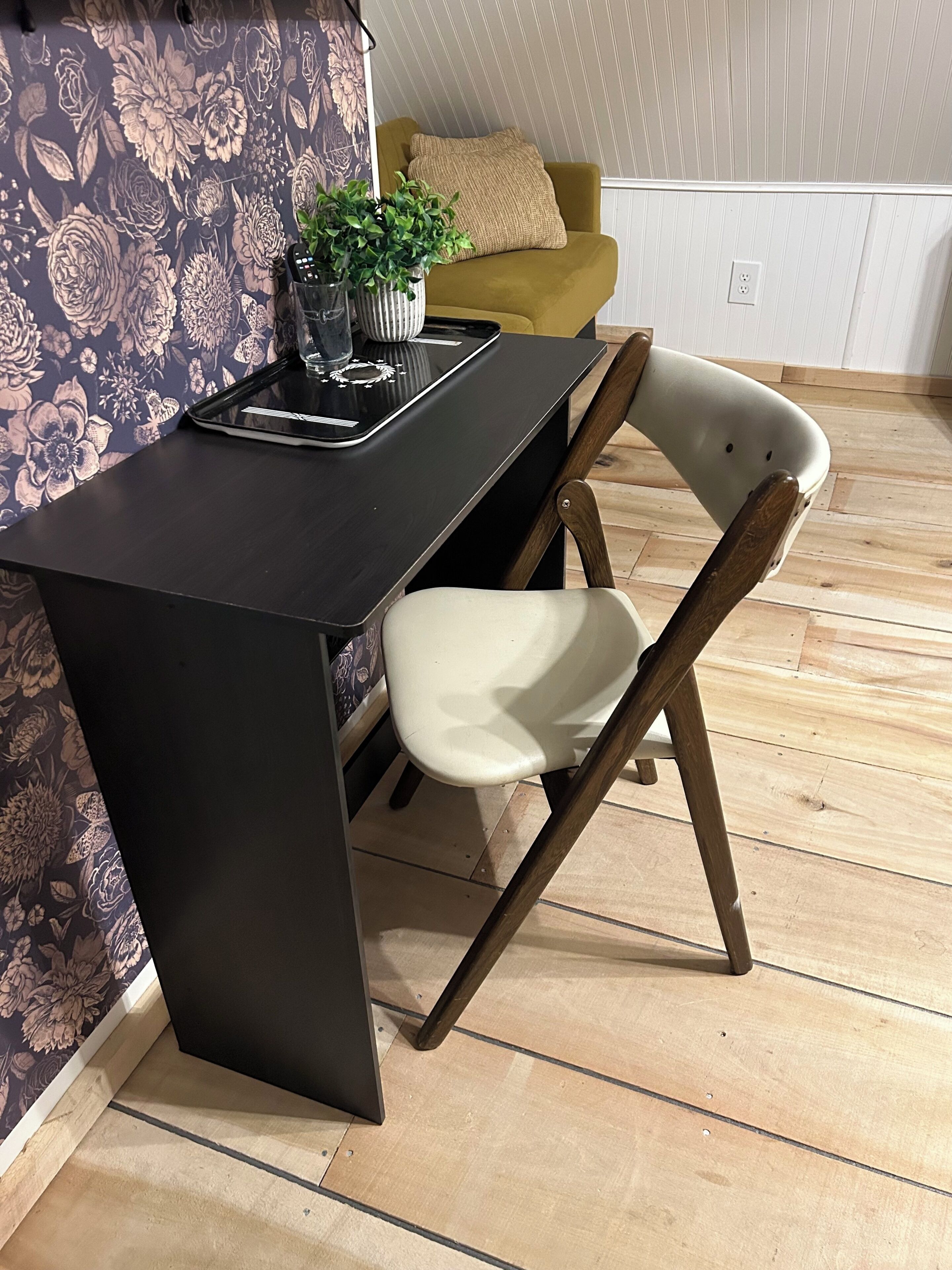 Una habitación con un escritorio negro, una silla blanca, un sofá amarillo y un papel tapiz con motivos florales.