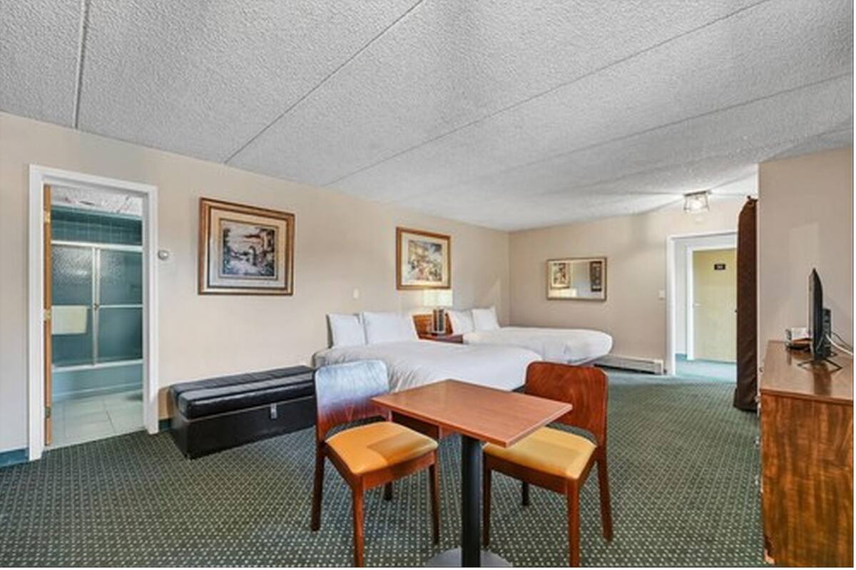 Une chambre d’hôtel comprenant un lit, un bureau avec deux chaises, un banc et deux tableaux encadrés accrochés au mur.