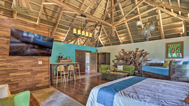 Un'ampia stanza con soffitto in legno a vista, un letto con copripiumino bianco e blu, una poltrona verde, un tavolino in legno e una TV a parete.