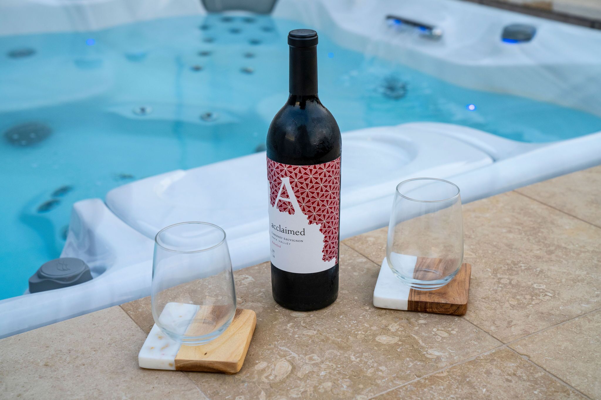 A bottle of wine and two glasses on a tiled surface near a hot tub.