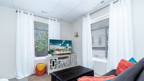 Ein modernes Wohnzimmer mit einem Flachbildfernseher, einem schwarzen Ledersitzhocker, einem Sofa mit bunten Kissen und einem kleinen weißen Tisch.