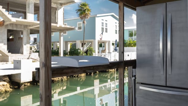 A modern kitchen with a stainless steel refrigerator, a wooden countertop, and a view of a marina with boats and a building in the background. - Anchor's Away: Discover Charm on the Canal, Swift Atlantic Access, Dockage & Above Ground Pool Await (Marathon)
