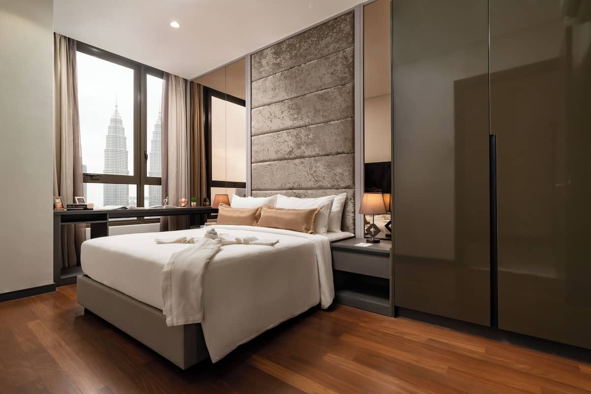 A modern hotel room with a large bed, a desk with a computer, a wardrobe, and a view of the city skyline.