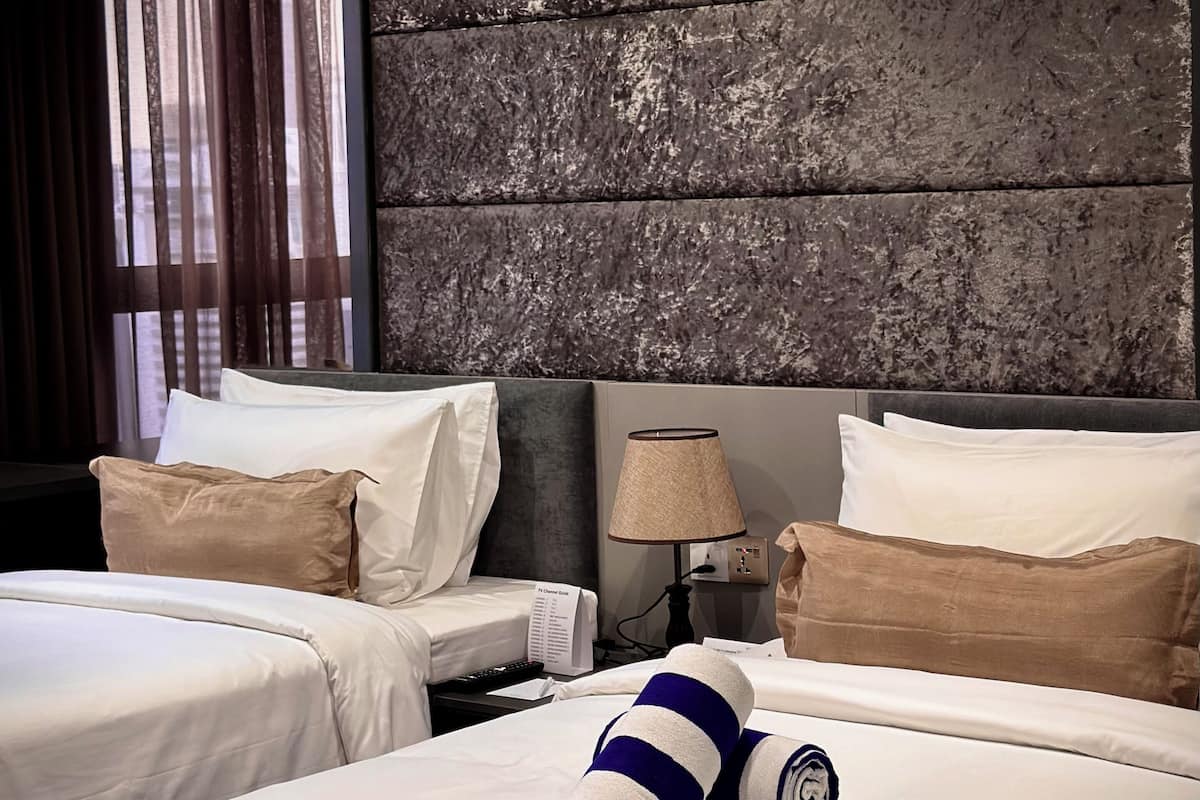 A hotel room with two beds, a textured wall, a lamp, and a view of the city through sheer curtains.