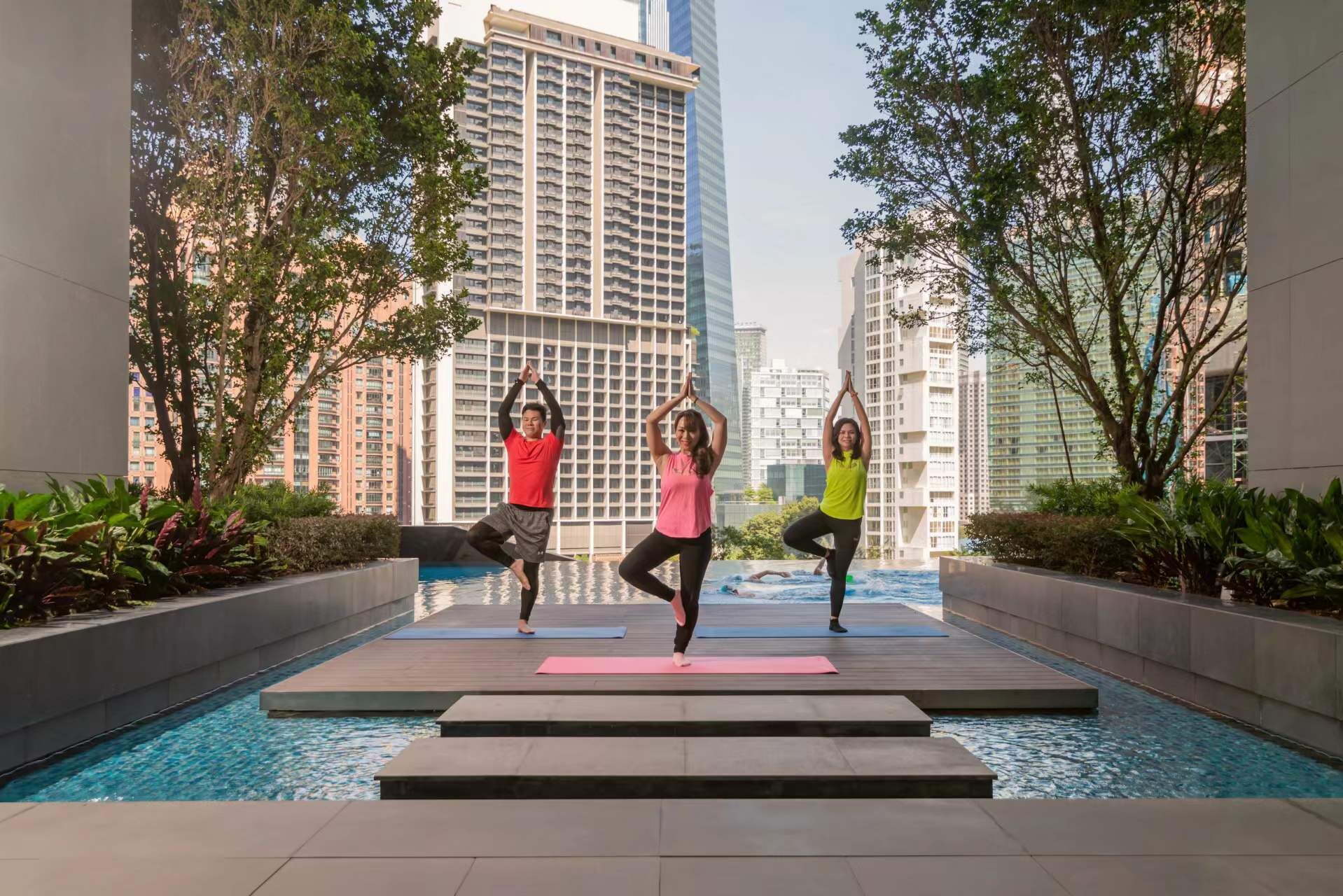 Two individuals performing yoga on a wooden platform by a pool with high-rise buildings in the background.