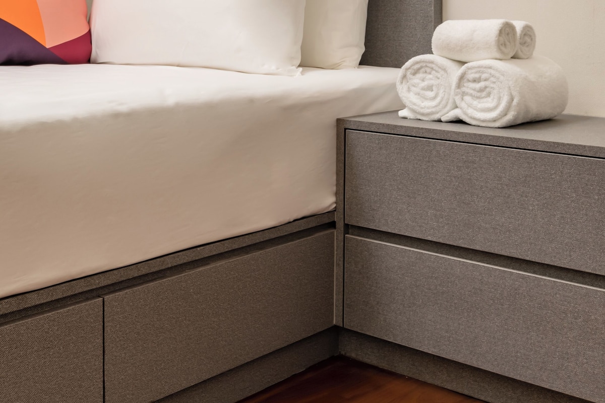 A neatly made bed with white linens and a pillow, a bedside table with rolled towels, and a colorful pillow.