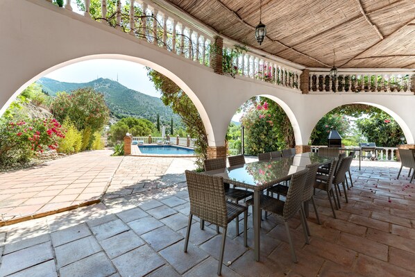 A covered patio with a glass table and wicker chairs, overlooking a swimming pool and lush greenery.