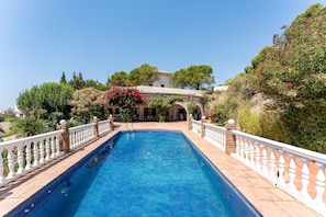 A rectangular swimming pool with a white balustrade, surrounded by greenery and a building with arches in the background.