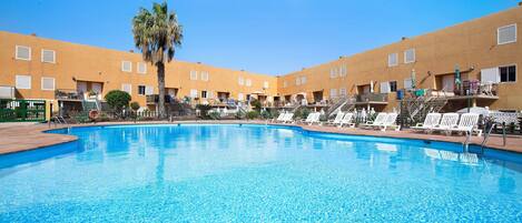 En swimmingpool med liggestole og en palme, omgivet af en bygning med altaner.