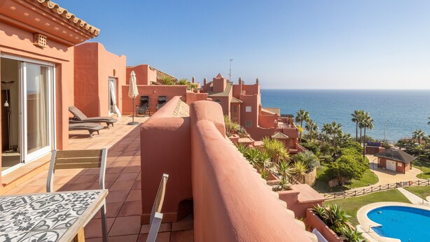 A balcony with a pool, a clear view of the sea, and a building with a terracotta facade.