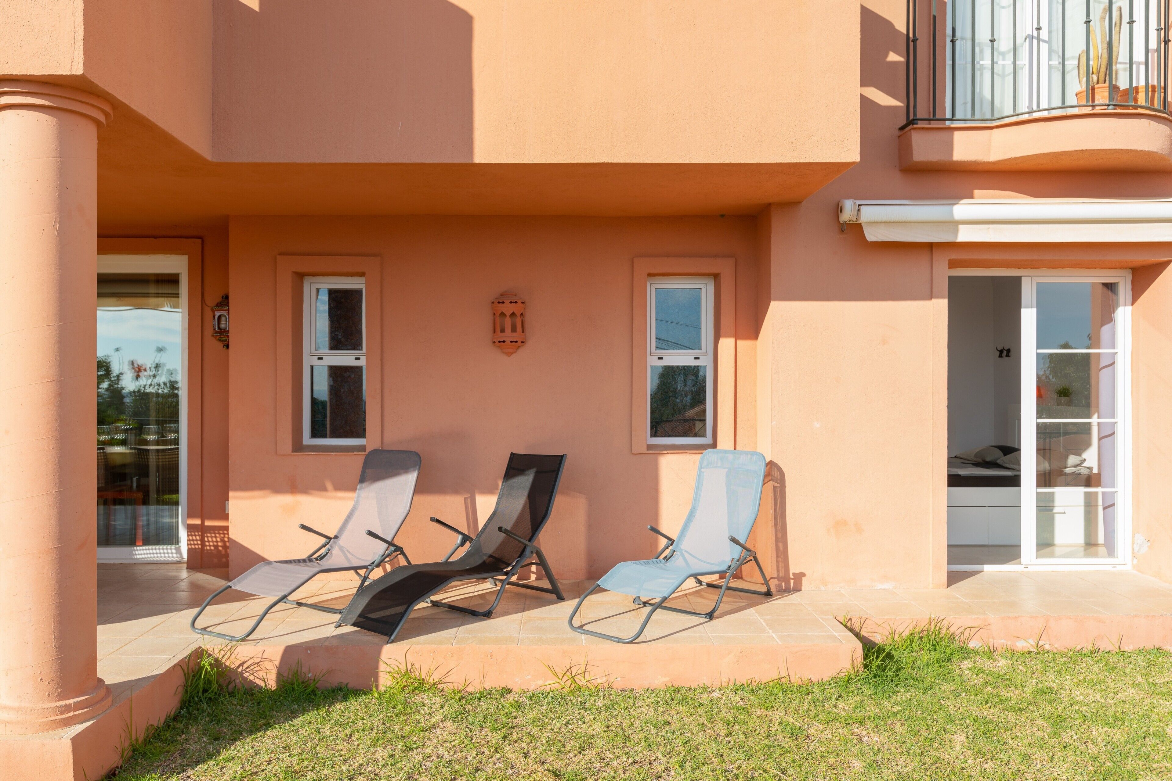 A building with a covered patio area, three lounge chairs, and a glass door entrance.