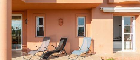 A building with a covered patio area, three lounge chairs, and a glass door entrance.