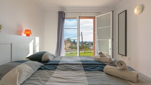Ein Schlafzimmer mit Bett, einem Fenster mit Vorhängen, einem Nachttisch mit Lampe und Blick ins Freie.