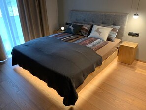 Une chambre à coucher moderne avec un lit, une table de chevet, une lampe et une vue sur l’extérieur.