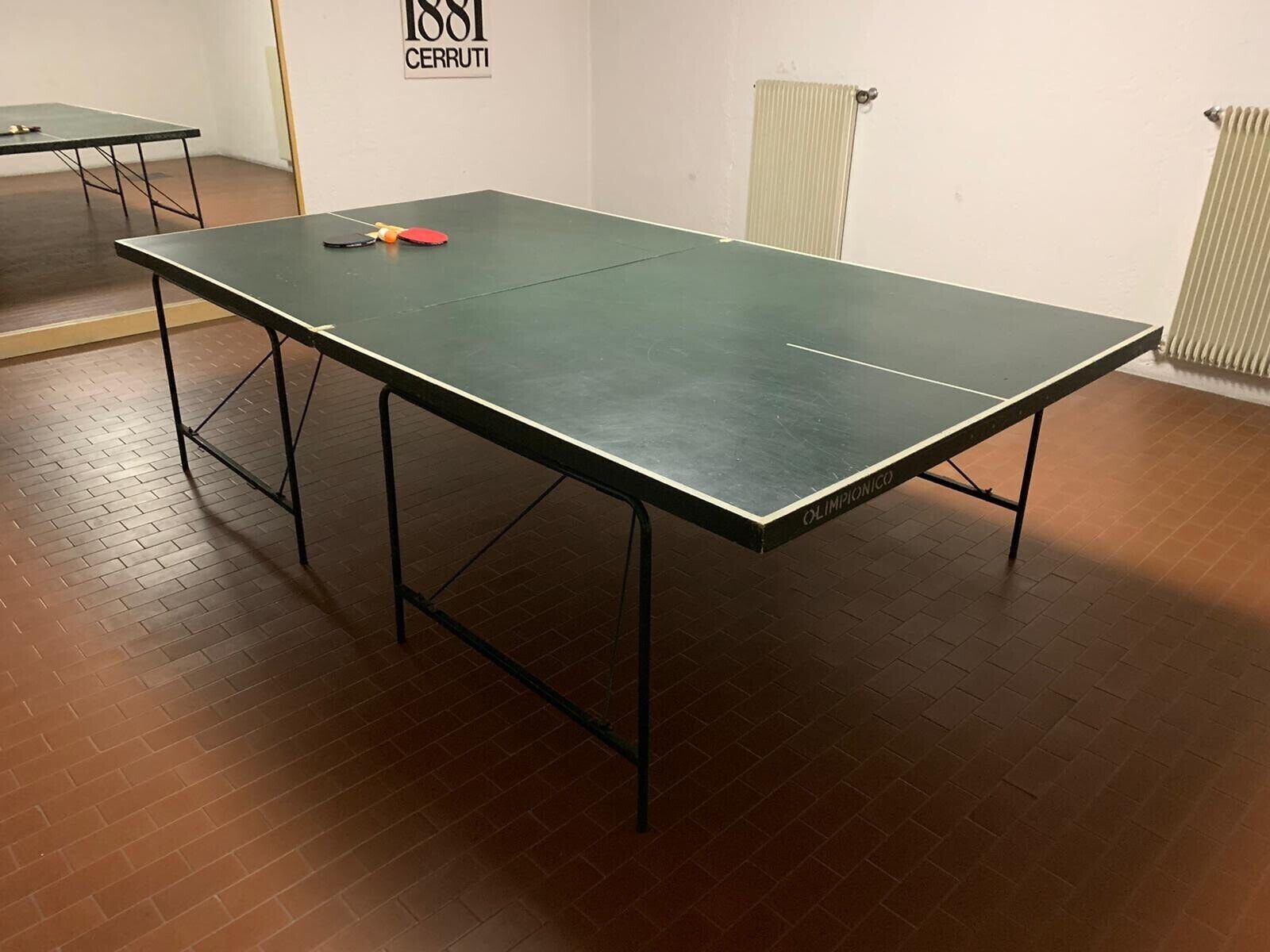 A ping pong table with a table tennis paddle and balls, a radiator, and a wall sign.