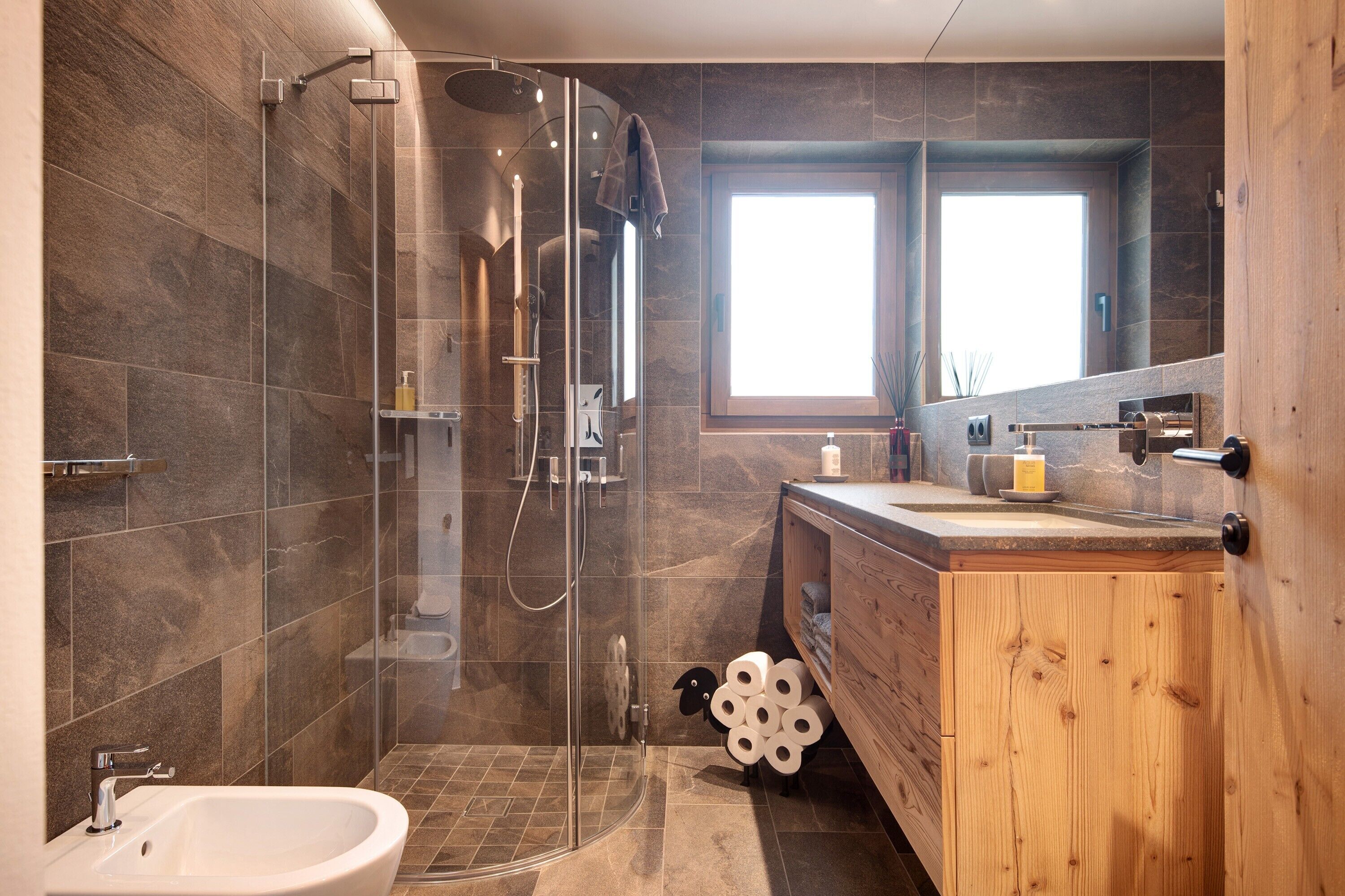 Une salle de bain moderne dotée d’une douche à parois de verre, d’un meuble-lavabo en bois et d’une grande fenêtre qui laisse entrer la lumière naturelle.