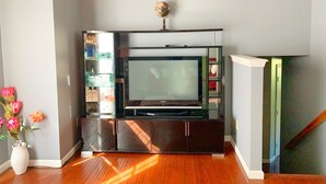 Un salon avec un téléviseur à écran plat posé sur un meuble noir, un parquet en bois et un escalier situé sur le côté droit.