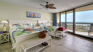 A hotel room with two beds, a ceiling fan, a painting, a bench, and a view of the ocean. - Coastal Calm | Split level condo with ocean views (Port Aransas)