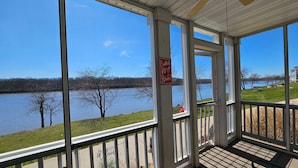 Eine überdachte Veranda mit großen Fenstern mit Blick auf einen Fluss, ein Schild, das auf Cocktails hinweist, und eine Aussicht auf eine Wiese sowie ein Gebäude gegenüber dem Wasser.