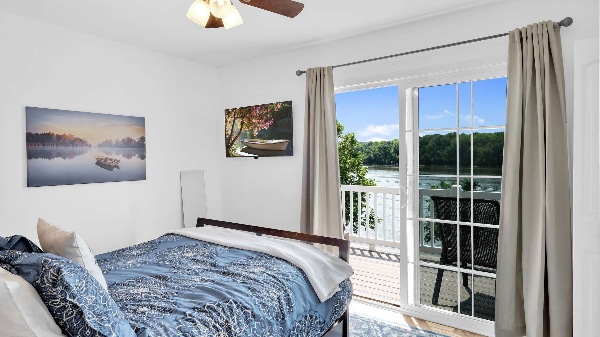 Una camera da letto con un letto, una ventola a soffitto, un quadro di un lago, una finestra con tende e un balcone con una sedia.