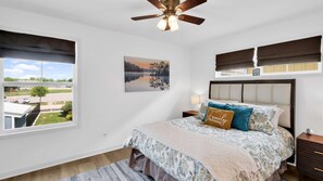 A bedroom with a bed, a nightstand, a lamp, a ceiling fan, a window with a view of a parking lot, and a painting on the wall.