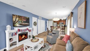 A living room with a fireplace, a television, a dining area, and a kitchen in the background.