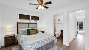 Un dormitorio con cama, dos mesitas de noche, ventilador de techo, ventana con persianas y vista al baño.