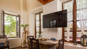 Un coin repas avec une table en bois, des chaises, un téléviseur fixé au mur et des rideaux en dentelle aux fenêtres.