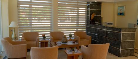 A lobby with beige armchairs, a reception desk, and a large window with blinds.
