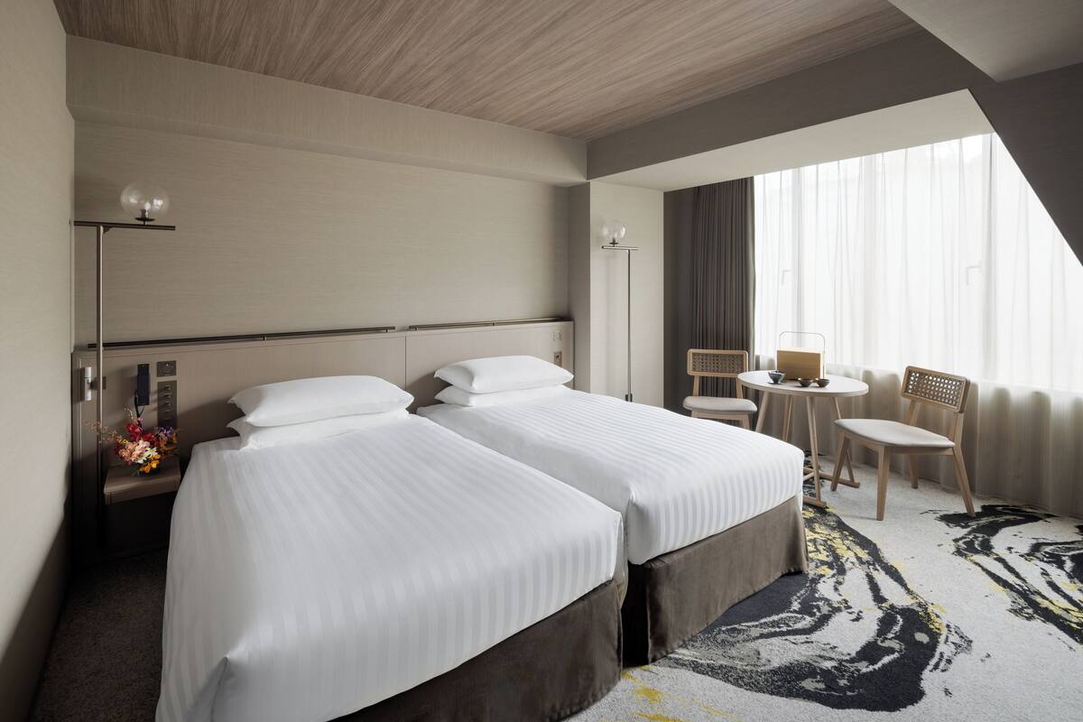 Ein Hotelzimmer mit zwei Betten, einem Schreibtisch, einem Stuhl, einem kleinen Tisch, einer Lampe und einem Fenster mit Vorhängen.
