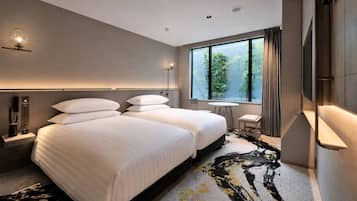 Ein Hotelzimmer mit zwei Betten, einem kleinen Tisch, einem Stuhl, einem Fenster mit Blick auf Grünflächen und einem modernen Design.