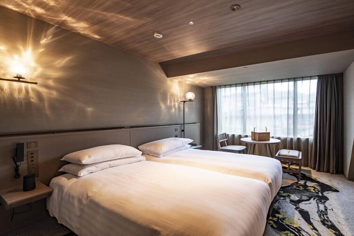 A hotel room with a large bed, bedside tables, a desk, a chair, and a view of the city through the window.