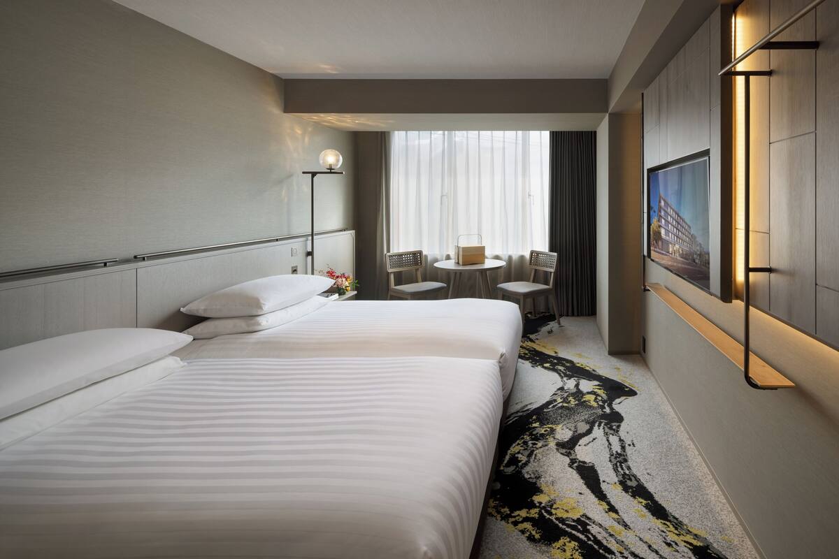 Ein Hotelzimmer mit zwei Betten, einem kleinen Tisch mit Stühlen, einem großen Fenster mit Vorhängen und einem an der Wand befestigten Fernseher.