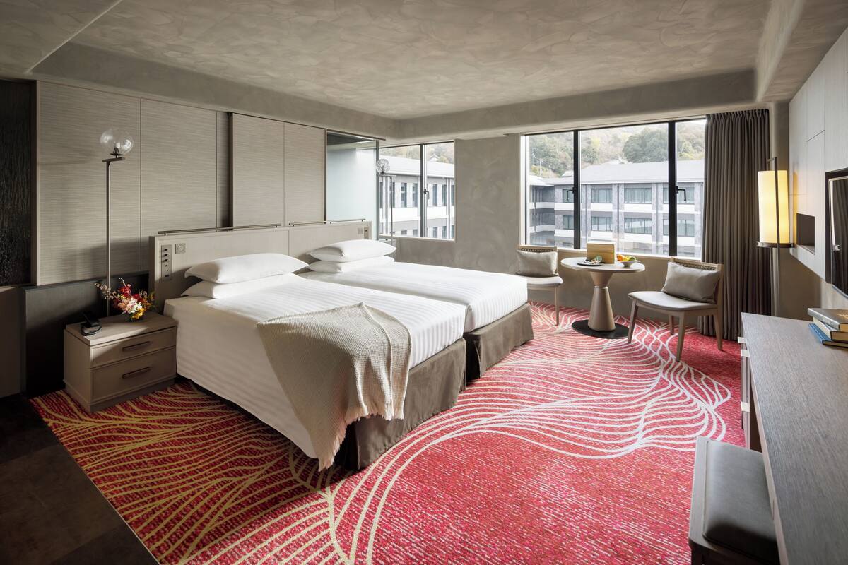 Ein Hotelzimmer mit einem großen Bett, Nachttischen, einem Schreibtisch, einem Sessel und Blick auf Gebäude durch das Fenster.